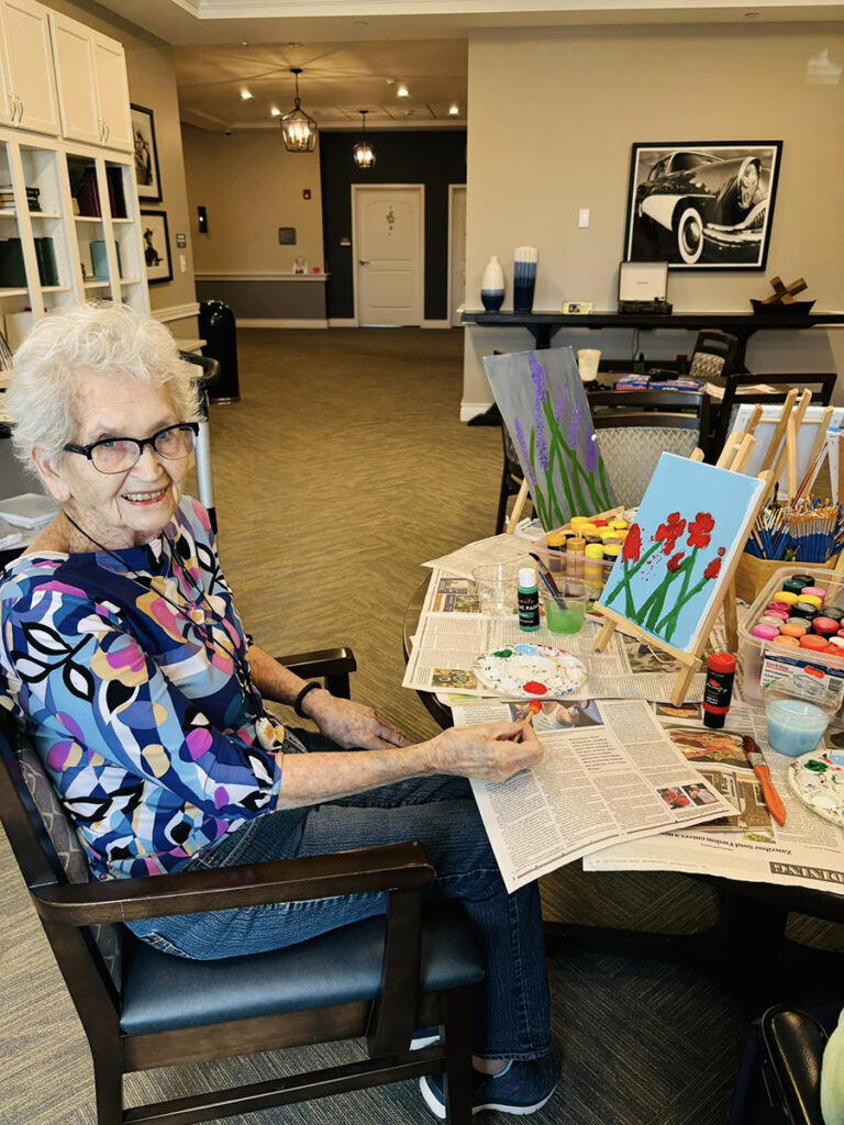 An elderly woman happily painting red flowers on a canvas at an arts and craft day in a senior living community.