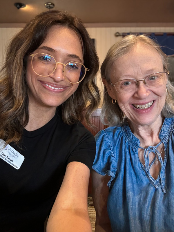 A senior resident and a staff member smile for a selfie. The senior is wearing a blue blouse, and the staff member is wearing glasses and a black shirt with a name tag.