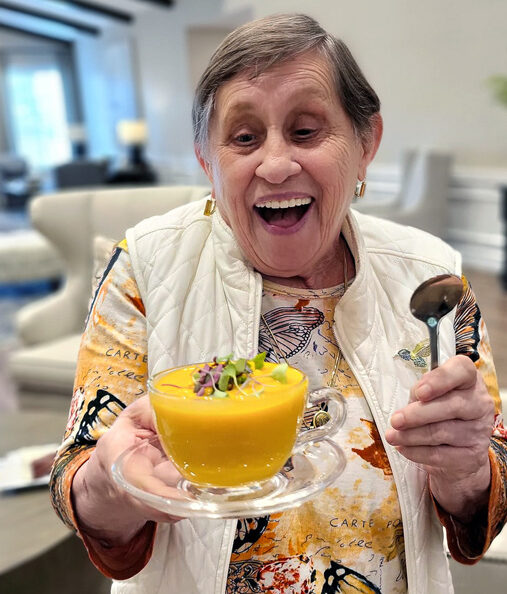 Smiling senior resident joyfully holds a bowl of vibrant orange soup garnished with greens, wearing a colorful butterfly-patterned shirt and white vest, ready to enjoy the meal.
