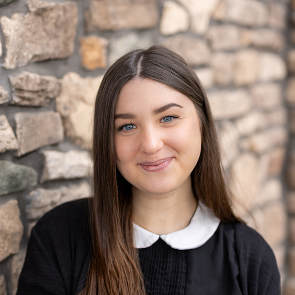 Isabella 'Bella' Hoeller, Business Office Director at Vitalia Rockside, smiles in front of a stone wall, wearing a black outfit with a white collared shirt.