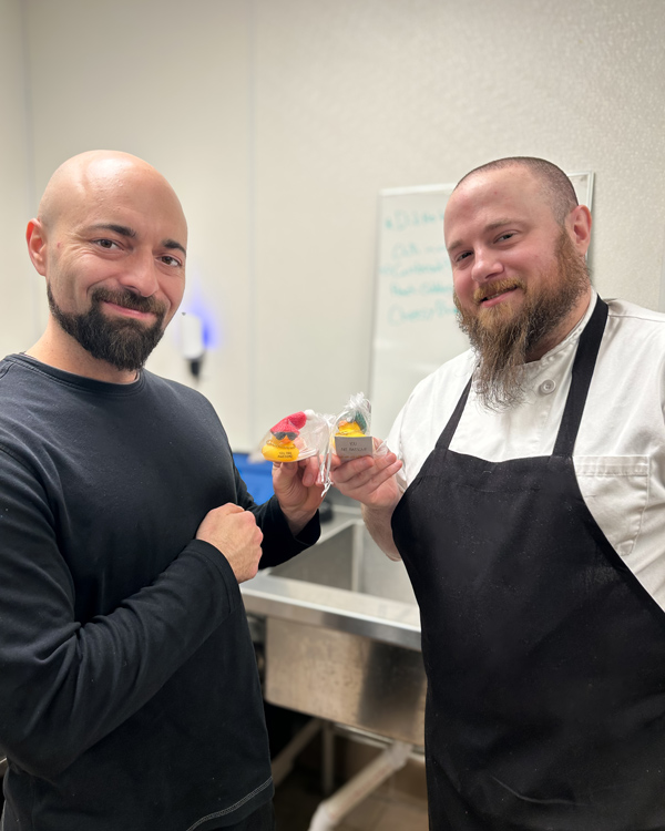 Two senior living culinary team members smile while holding small appreciation gifts wrapped with festive ribbons, celebrating Employee Appreciation Day in the kitchen.