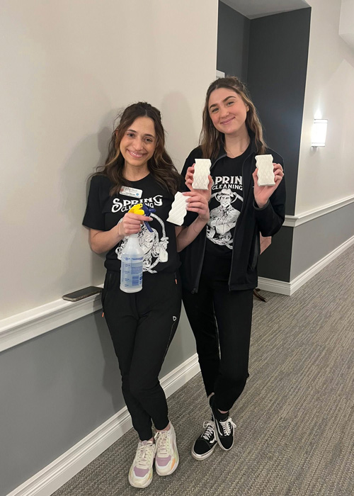 Two women in matching spring cleaning shirts smiling while holding spray bottle and scrub pads.