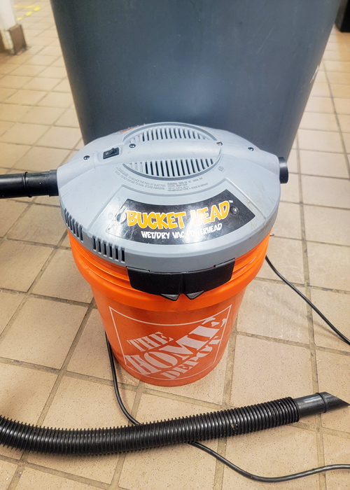 Bucket Head wet/dry vacuum attached to an orange Home Depot bucket, sitting on a tiled floor in a commercial or industrial cleaning area.
