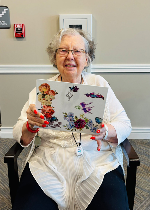 Smiling senior woman seated in a chair holding a white board decorated with colorful flower and bird stickers at a senior living community.