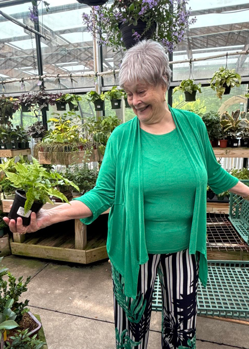 Senior woman in a green outfit smiles while holding small potted plants in a vibrant greenhouse setting.