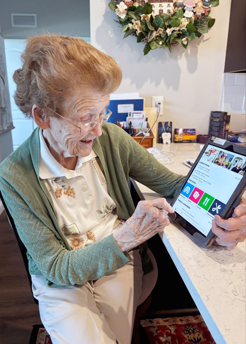 A smiling senior resident uses a tablet to explore features of the Arrow app while seated at a kitchen counter, engaging with technology independently.