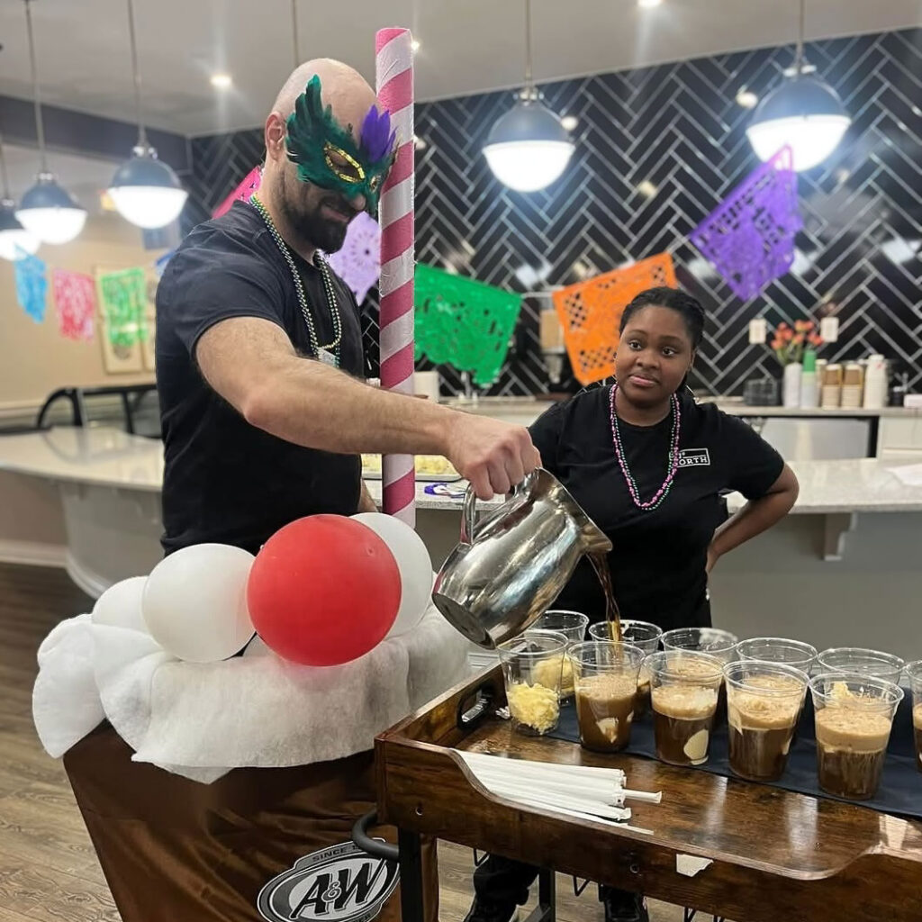 A Culinary Director in a Mardi Gras mask pours root beer floats into cups from a festive cart, as a team member watches during a lively celebration in the community.