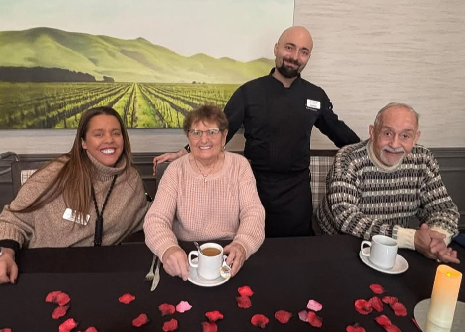A Culinary Director stands with two senior residents and a team member at a decorated dining table, sharing coffee and smiles during a warm community gathering.