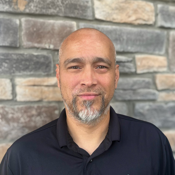 Hector Santiago, Plant Operations Director at Vitalia Rockside, smiling in a black collared shirt in front of a stone wall background.