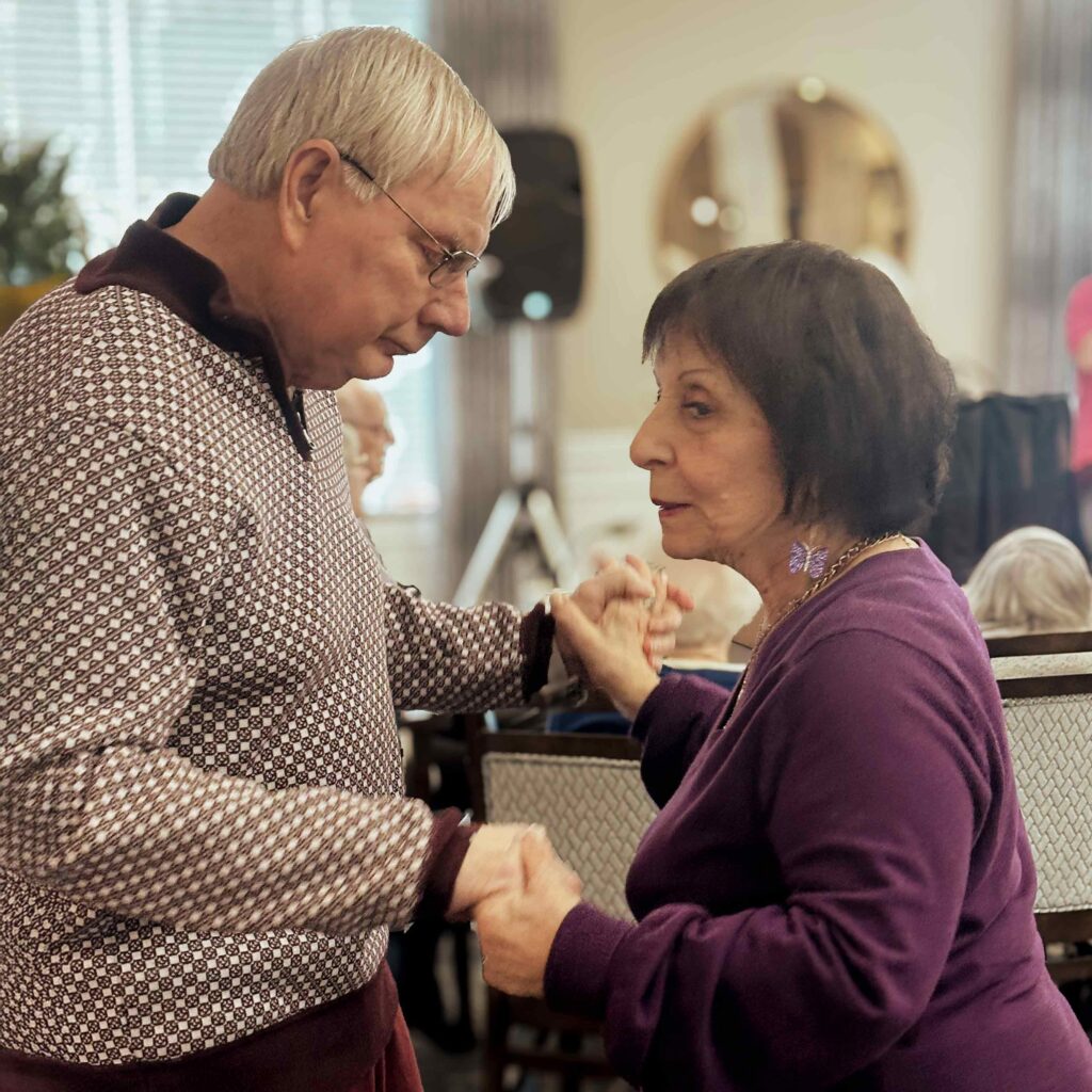 Senior couple holding hands while dancing together in a community space, sharing a warm and joyful moment during a group event.