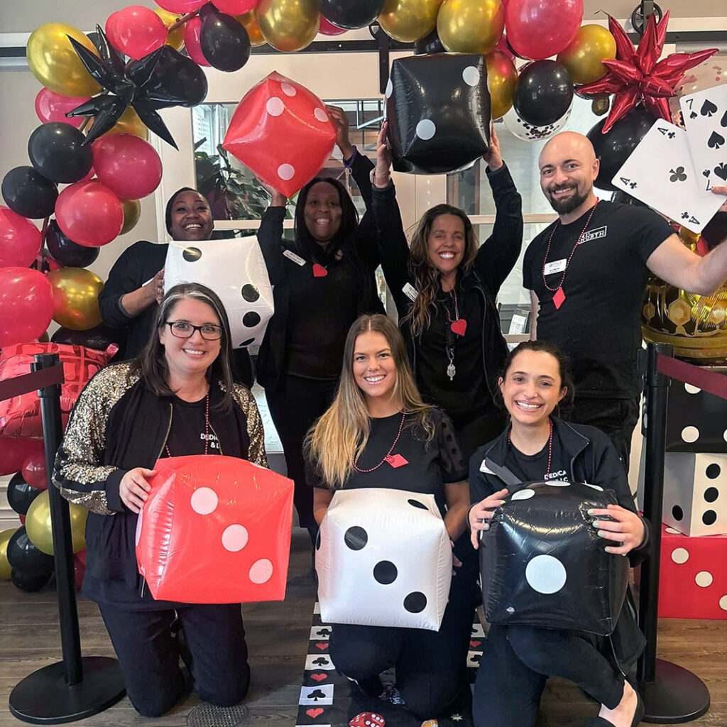 Team members smile under a balloon arch, holding large inflatable dice during a fun event.