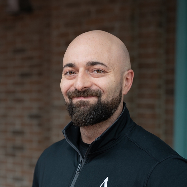 Alexander “Alex” Bratsch, Culinary Director at Vitalia Rockside, smiling in a professional headshot, wearing a black zip-up jacket, with a softly blurred brick background.