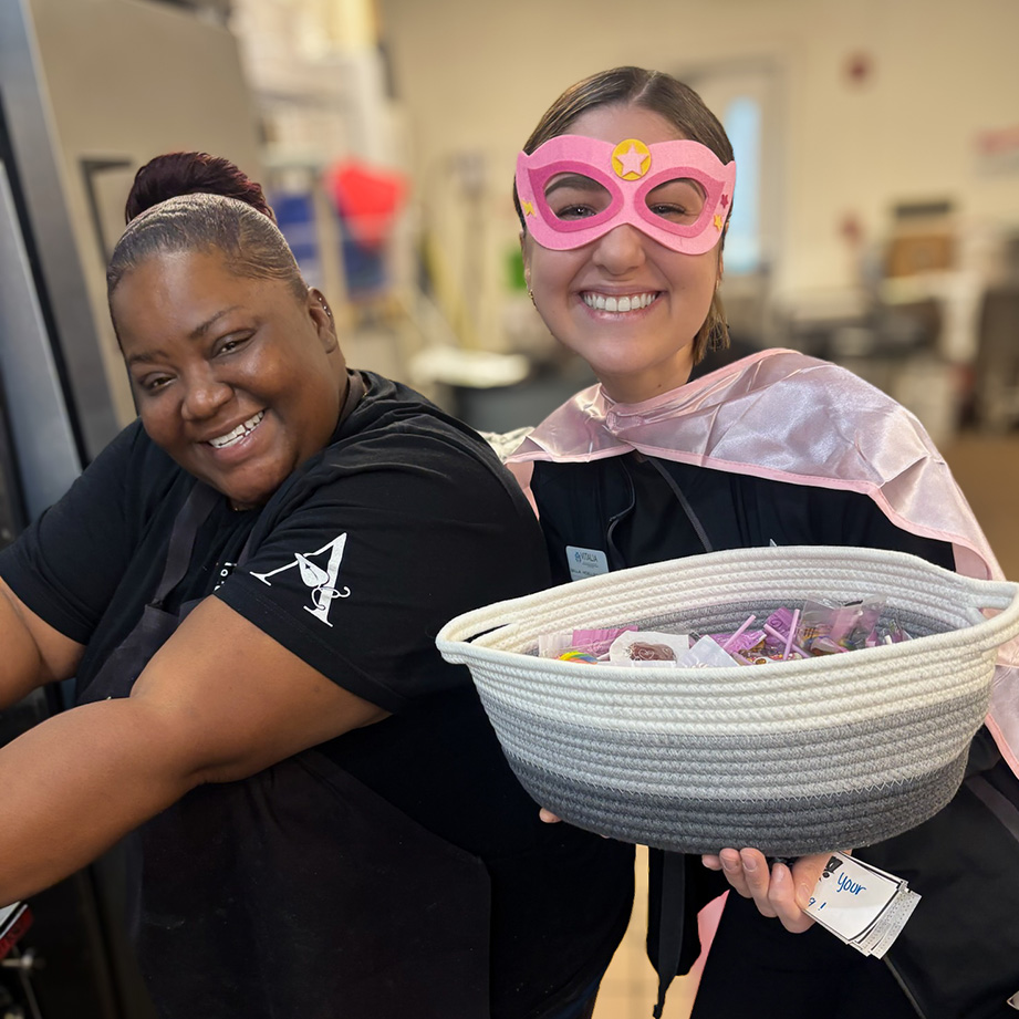 Two women on the Vitalia Rockside team smile brightly, one wearing a pink superhero costume, passing out treats.