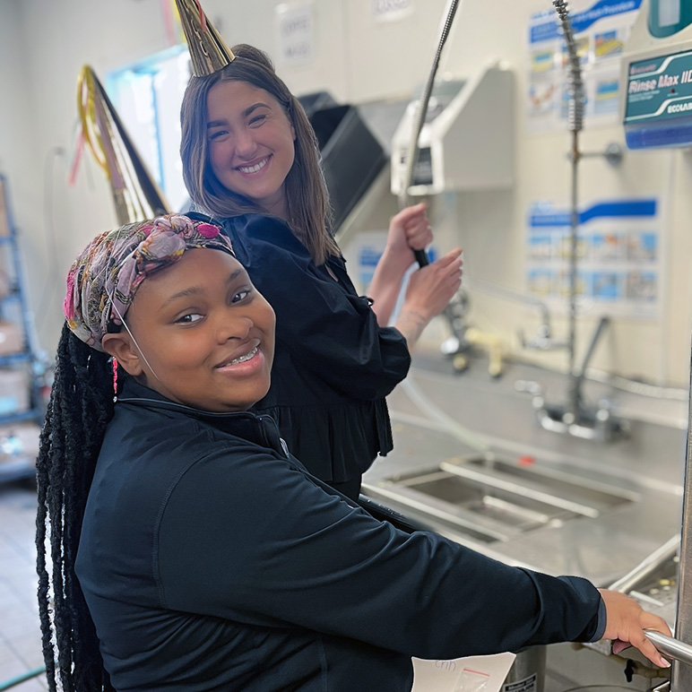 Two women wearing party hats smile while doing a deep cleaning procedure in the Vitalia Rockside kitchen.