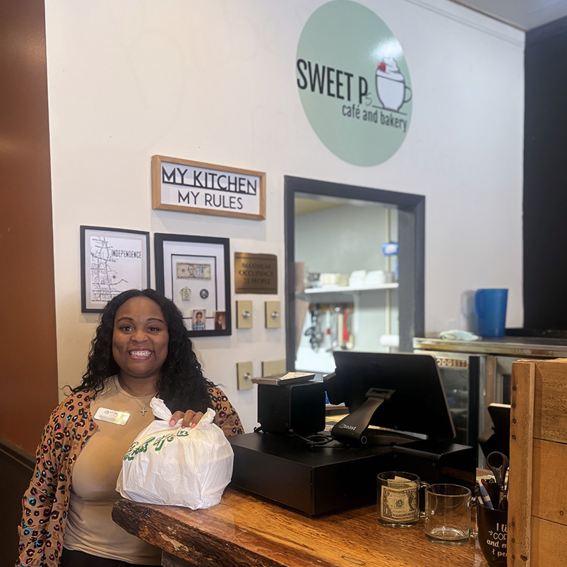 A woman on the Vitalia Rockside team smiles brightly at Sweet P's Cafe and Bakery, holding a to-go bag.
