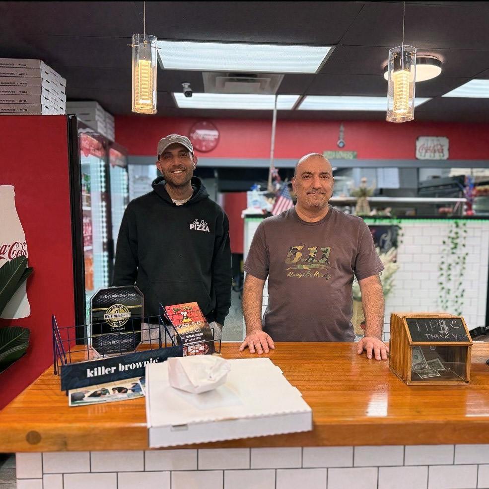 Two men smile proudly behind the counter of their establishment during a visit from Vitalia Rockside.