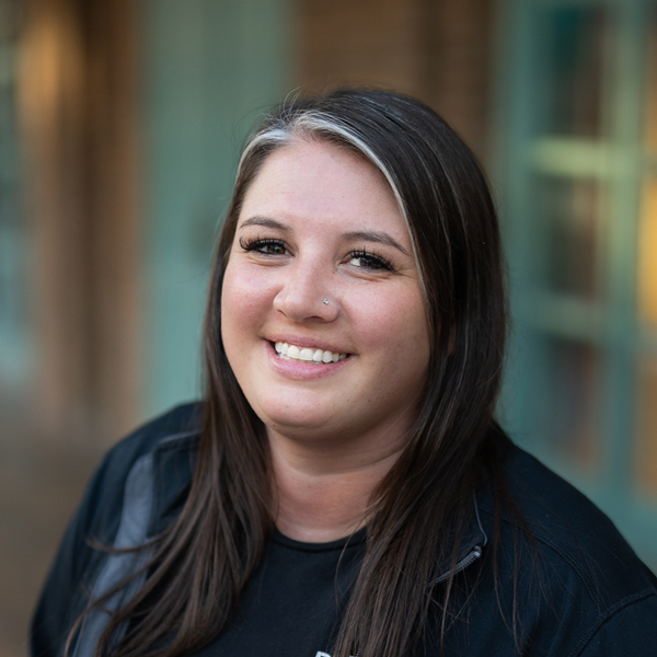 Stephanie Sather, Executive Director at Vitalia Rockside, smiling in a professional outdoor headshot with long dark hair, a light front streak, and softly blurred brick and teal window details in the background.