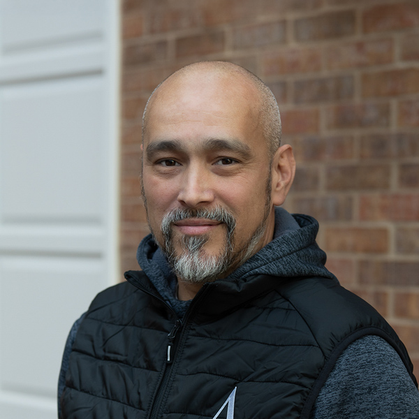 Hector Santiago, Plant Operations Director at Vitalia Rockside, looking at the camera in a professional outdoor headshot turned slightly to the side, with a bald head, a salt-and-pepper goatee, a black vest over a dark hoodie, and a softly blurred brick background.