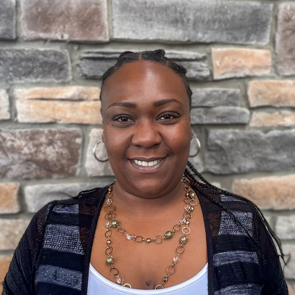 Jovonn Gaskin, Senior Living Director at Vitalia Rockside, smiling in a professional headshot with braided hair pulled back, hoop earrings, layered gold necklaces, and a stacked stone wall background.