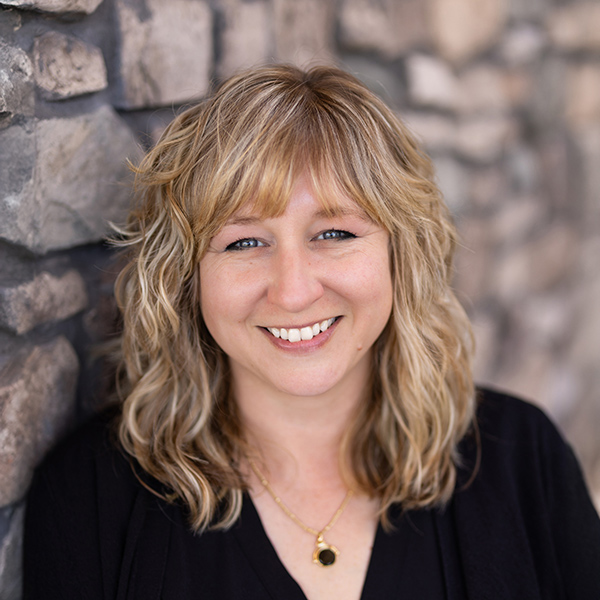 Angela Raresheid, Resident Services Director at Vitalia Rockside, smiles in a professional headshot with wavy blonde hair, a black top, and a stone wall background.
