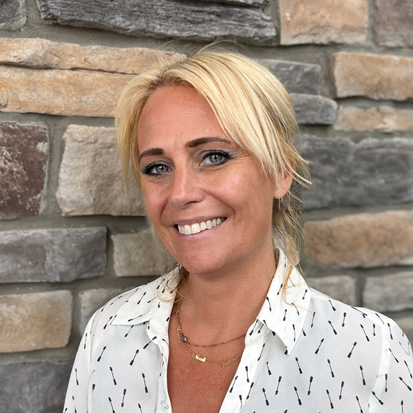 Lindsay Shamaly, Memory Care Director at Vitalia Rockside, smiling in a professional headshot with blonde hair pulled back, a white patterned blouse, layered necklaces, and a stacked stone wall background.