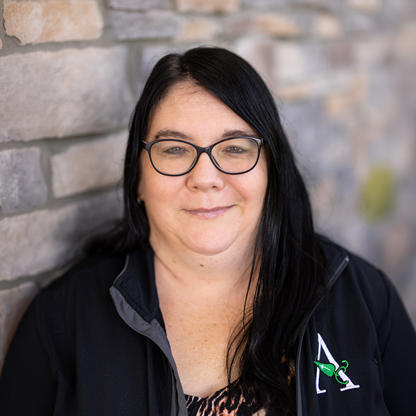 Andrea Mulhall, Operations Coordinator at Vitalia Rockside, looking at the camera in a professional headshot with long dark hair, black glasses, a black jacket, and a softly blurred stone wall background.