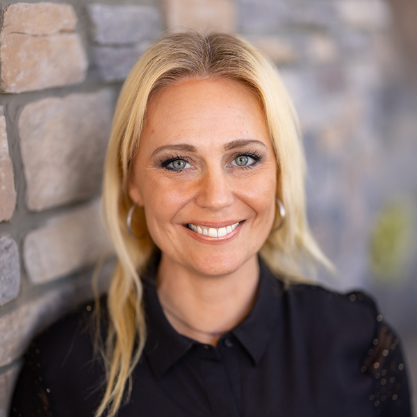 Lindsay Shamaly, Memory Care Director at Vitalia Rockside, smiling in a professional headshot with blonde hair, gold hoop earrings, a black blouse, and a softly blurred stone wall background.